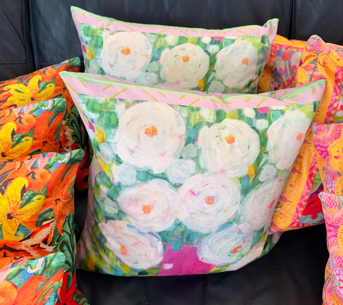 Two decorative throw pillows with a colorful floral and botanical print are arranged on a black leather couch, alongside other patterned cushions.